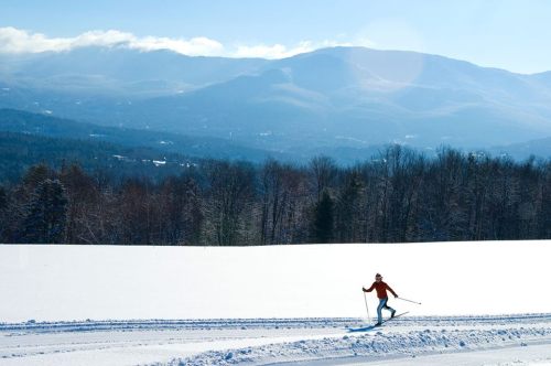 A skier glides across a snowy landscape with mountains in the background under a clear blue sky.