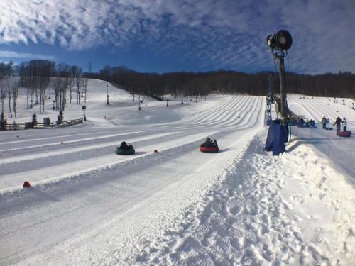 A snowy hill with people tubing down, surrounded by trees and a clear blue sky. Snowmaking equipment in the background.