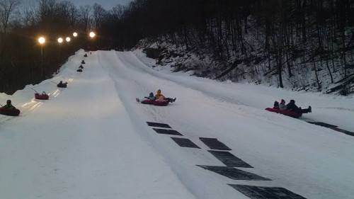 People tubing down a snowy slope at night, with lights illuminating the hill and trees in the background.