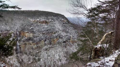 Snow-covered cliffs and trees in a winter landscape, with a waterfall visible in the distance.