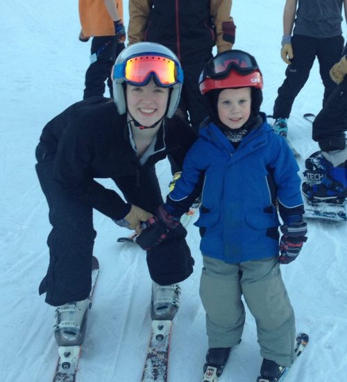 A smiling young woman and a boy in ski gear pose together on a snowy slope, surrounded by other skiers.
