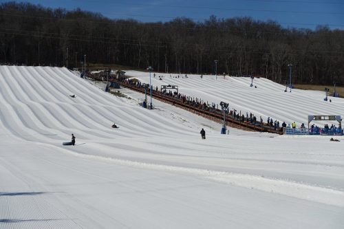 A snowy hill with multiple tubing lanes and a crowd of people waiting in line for their turn to slide down.