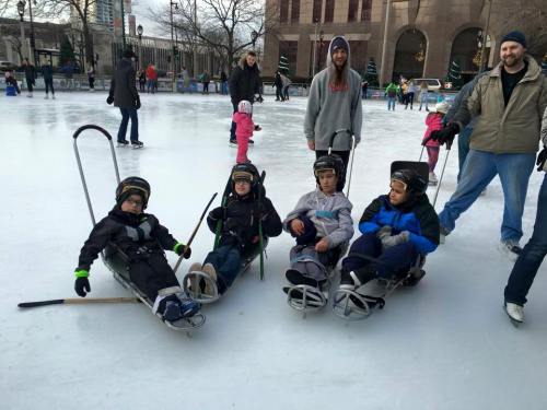 Four children in sleds on an ice rink, wearing helmets, with adults and other skaters in the background.