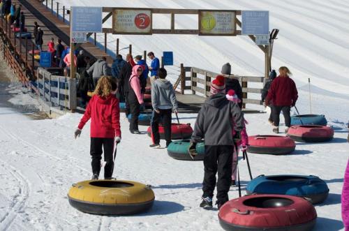 People walking on snow with colorful snow tubes, heading towards a tubing hill with a sign indicating lanes.