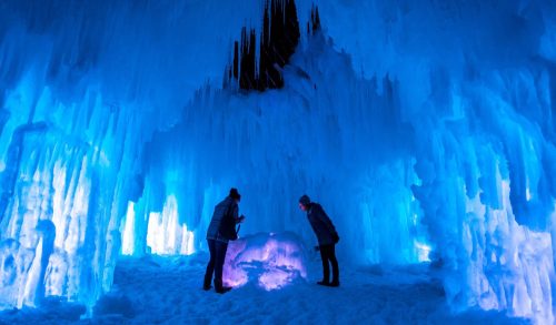 Two people explore a stunning ice cave illuminated by blue lights, surrounded by towering ice formations.
