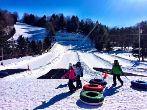 A snowy hill with people walking up, carrying colorful snow tubes, and others sliding down in the background.