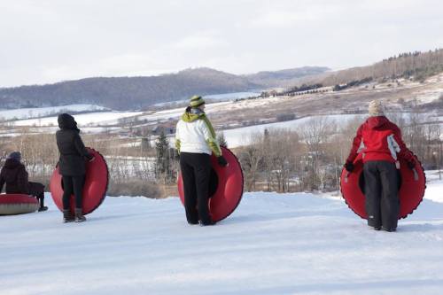 Four people stand on a snowy hill, each holding a red snow tube, with a scenic winter landscape in the background.