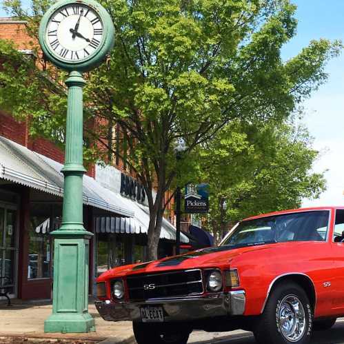 A vintage red car parked beside a green clock tower, with trees and shops in the background.