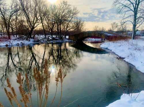 A serene winter scene featuring a stone bridge over a calm river, surrounded by snow-covered trees and grasses.