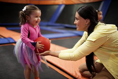 A smiling girl in a pink outfit holds a red ball while a woman in a yellow top reaches out to her in a trampoline park.