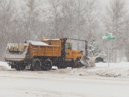 A snowplow truck clears snow on a winter road, with trees and a street sign visible in a snowy landscape.