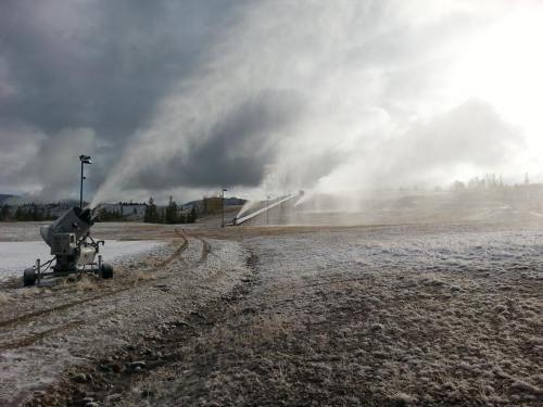 A snow-making machine sprays artificial snow onto a snowy field under a cloudy sky.