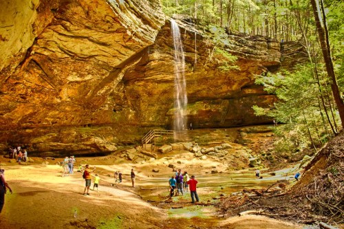 A scenic waterfall cascades down rocky cliffs, surrounded by lush trees and people exploring the sandy area below.