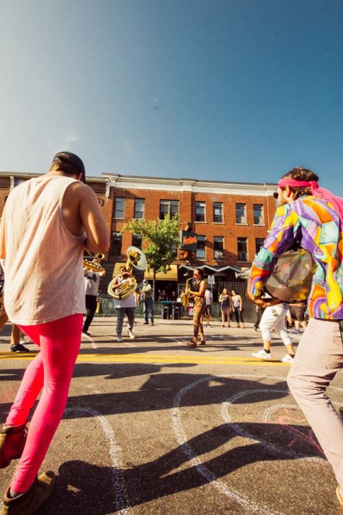 A lively street scene with musicians and dancers, colorful outfits, and a festive atmosphere under a clear blue sky.