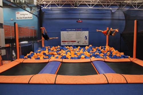 Two people performing flips above a foam pit filled with blue and orange blocks at a trampoline park.