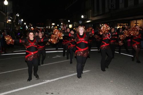 A group of dancers in festive outfits with pom-poms perform in a nighttime parade on a city street.