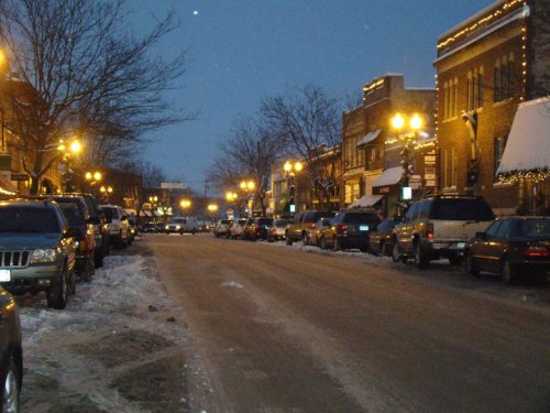 A snowy street lined with parked cars and illuminated buildings, creating a cozy evening atmosphere.