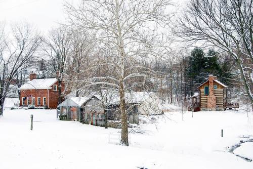 A snowy landscape featuring a brick house, a wooden cabin, and bare trees in a serene rural setting.