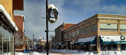 Snow-covered street with shops, festive wreaths, and a clear blue sky in a small town during winter.