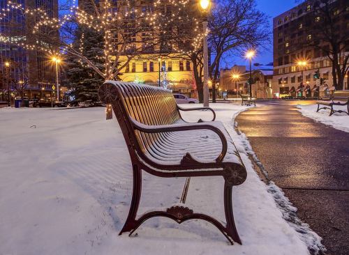 A snow-covered park bench illuminated by streetlights, with festive lights in the background at dusk.