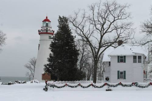 A snowy landscape featuring a lighthouse and a white house, both decorated with festive garlands and surrounded by bare trees.