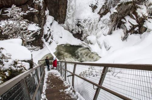 Two people stand on a snowy staircase overlooking a frozen waterfall and icy pool in a winter landscape.
