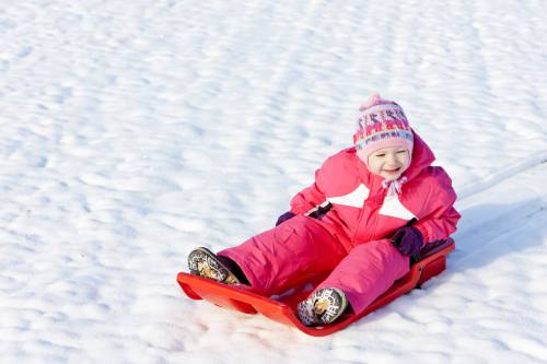 A smiling child in a pink snowsuit sits on a red sled in a snowy landscape.