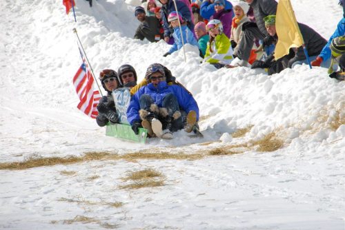 A group of people sledding down a snowy hill, with spectators cheering on the sides, some holding flags.