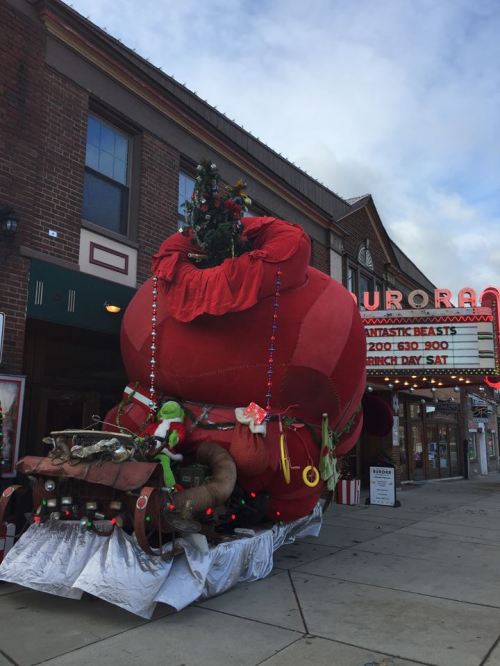 A large, festive Grinch-themed decoration with a red sack and Christmas tree outside a brick building.