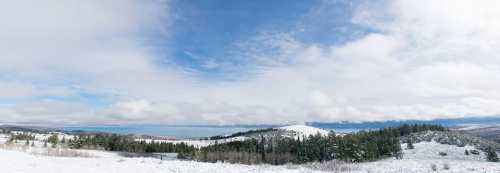 A panoramic winter landscape featuring snow-covered hills, evergreen trees, and a cloudy blue sky.