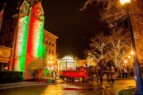 A festive night scene featuring a clock tower, illuminated trees, and a horse-drawn carriage in a lively town square.