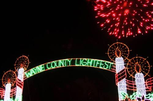 Colorful holiday lights form an arch reading "Wayne County Lightfest," with fireworks in the night sky above.