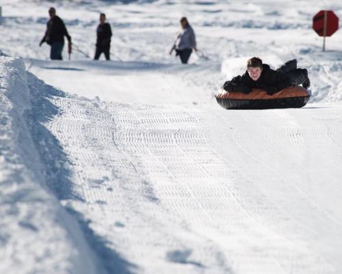 A person sleds down a snowy hill on a tube, while others walk in the background.