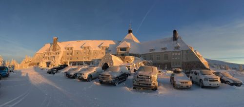 A snow-covered lodge with vehicles parked in front, surrounded by a winter landscape under a clear blue sky.