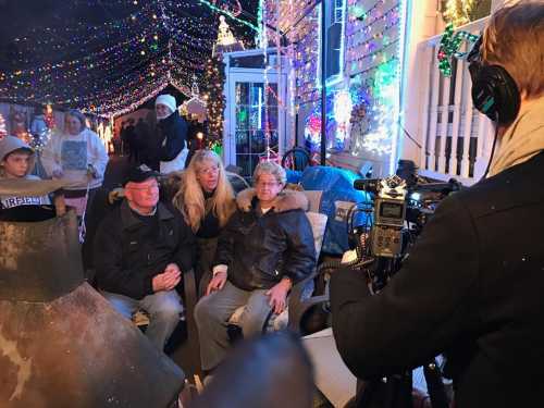 A group of people sits together in a festive, brightly lit outdoor setting, while someone films them.