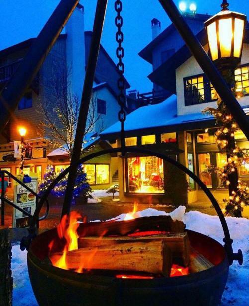 A cozy outdoor fire pit glows warmly in a snowy village, with festive lights and buildings in the background.