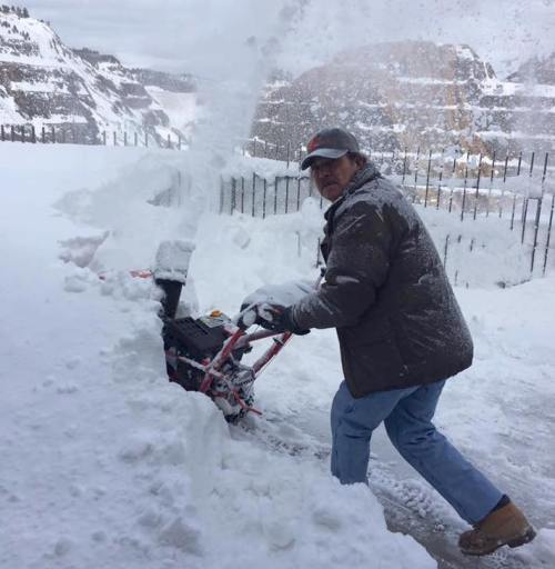 A man shovels snow with a snowblower in a snowy landscape, surrounded by tall snowbanks and a mountainous backdrop.