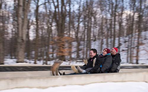 Three people joyfully sliding down a snowy track, surrounded by trees, with a sense of speed and excitement.