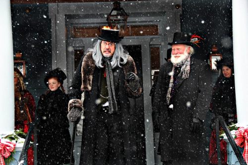 Two men in top hats and winter coats stand outside in the snow, surrounded by people in festive attire.