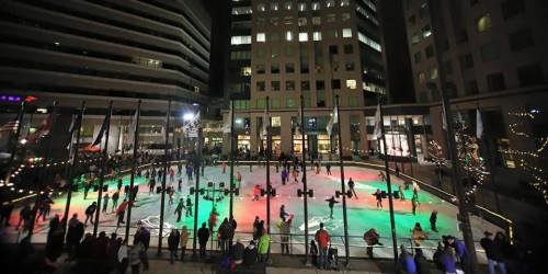 A bustling outdoor ice skating rink at night, illuminated with colorful lights and surrounded by tall buildings.