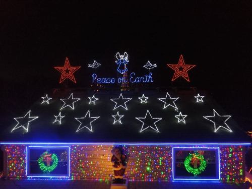 A house decorated with colorful Christmas lights, featuring stars, wreaths, and the phrase "Peace on Earth."