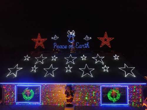 A house decorated with colorful Christmas lights, featuring stars, wreaths, and the phrase "Peace on Earth."