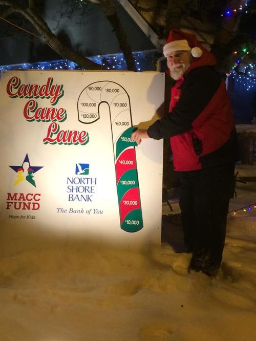 A man in a Santa outfit stands next to a fundraising thermometer sign for Candy Cane Lane, showing progress towards a goal.