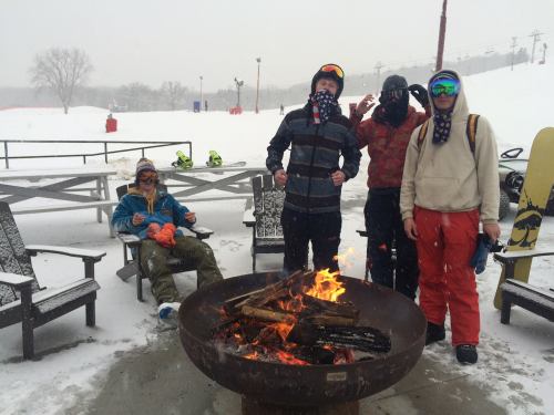Group of four people in winter gear around a fire pit, snow falling, with ski equipment in the background.