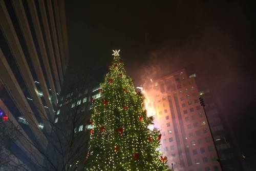 A brightly lit Christmas tree stands tall amidst city buildings, with smoke rising from a nearby structure.