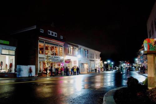 A festive street scene at night, with shops adorned in lights and reflections on wet pavement.