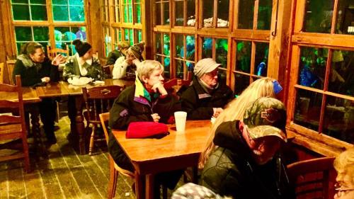 A cozy restaurant interior with people seated at wooden tables, enjoying food and drinks, surrounded by large windows.