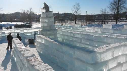 A snowy landscape with people building an ice structure, featuring stacked ice blocks and a person on top.