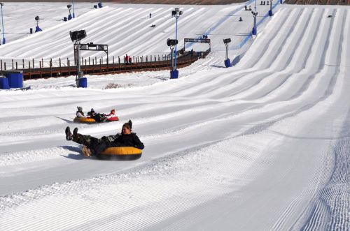 People tubing down a snowy slope, with groomed tracks and blue barriers in a winter landscape.