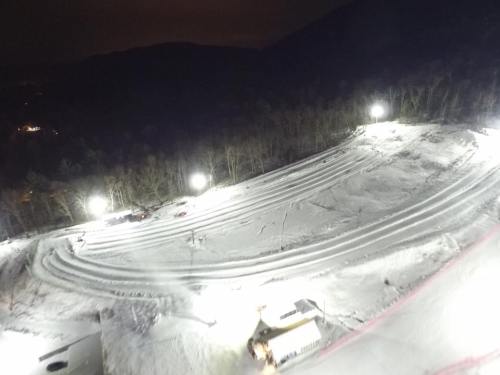 Aerial view of a snow-covered slope at night, illuminated by bright lights, with tire tracks visible in the snow.